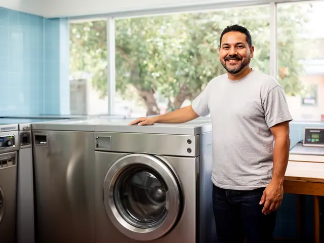 Laundromat owner in a grey t-shirt resting a hand on a large stainless-steel front-loading washer